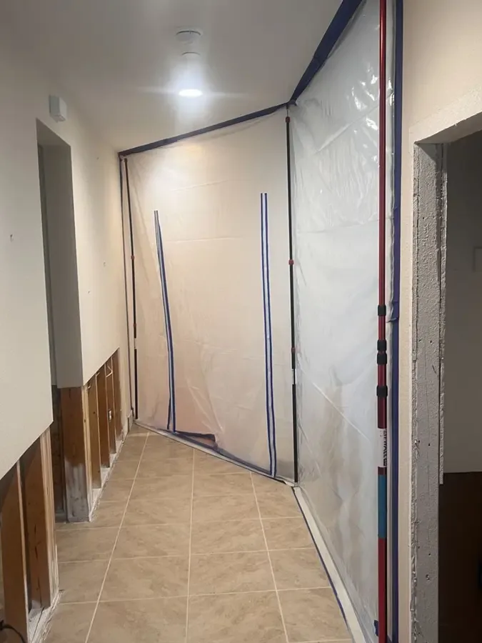 A hallway has removed lower drywall and plastic containment surrounding an interior doorway